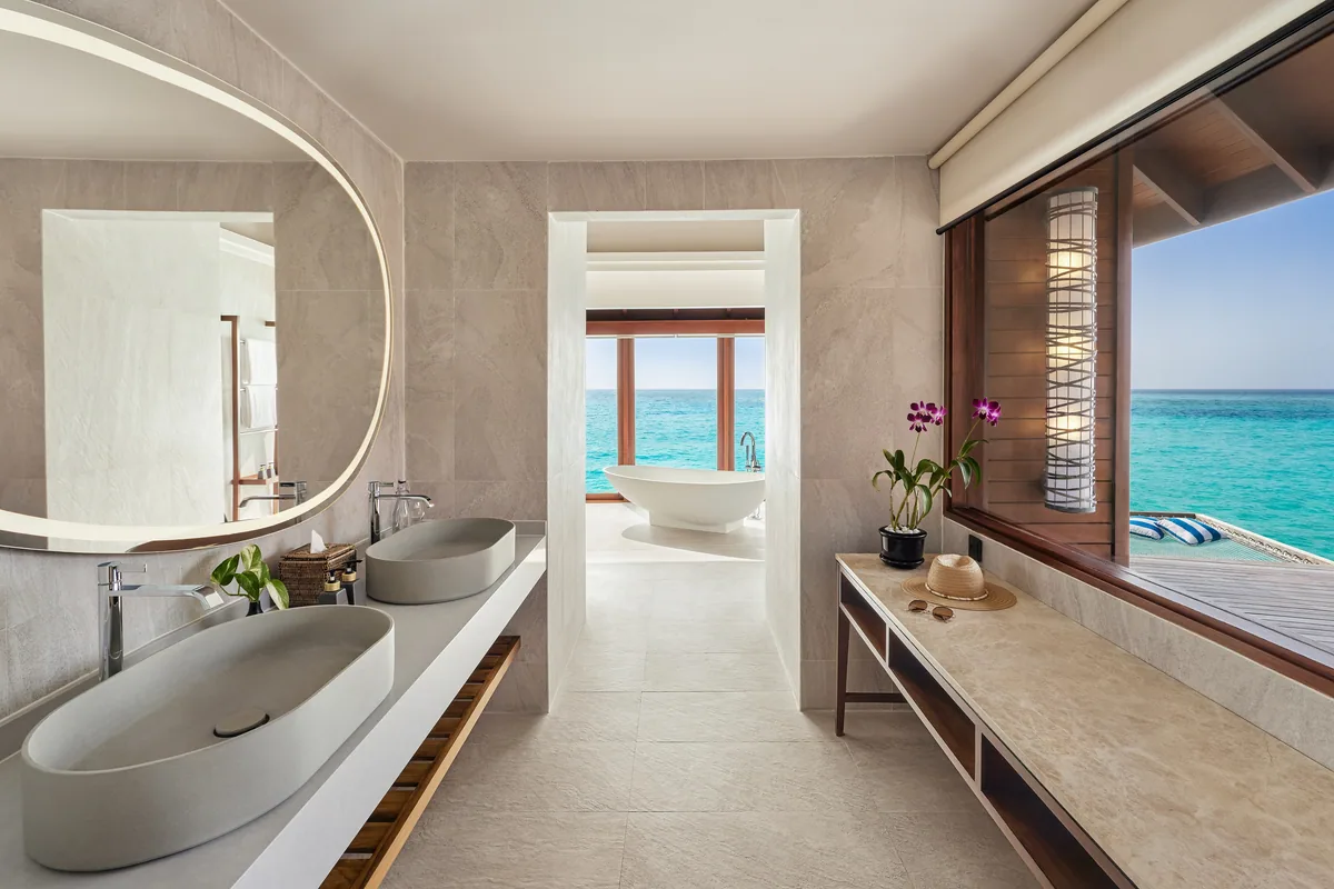 Anantara Dhigu Maldives Resort, villa overwater, South Malé Atoll, Maldives, Sunset Over-Water Villa bathroom, the twin grey-stone vessel sinks on a long stone counter, the large oval pill-shape vanity mirror, the freestanding white sculptural bathtub through the doorway with the open ocean horizon as backdrop, a magenta orchid on the side counter, and the over-water hammock visible through the right-side window.