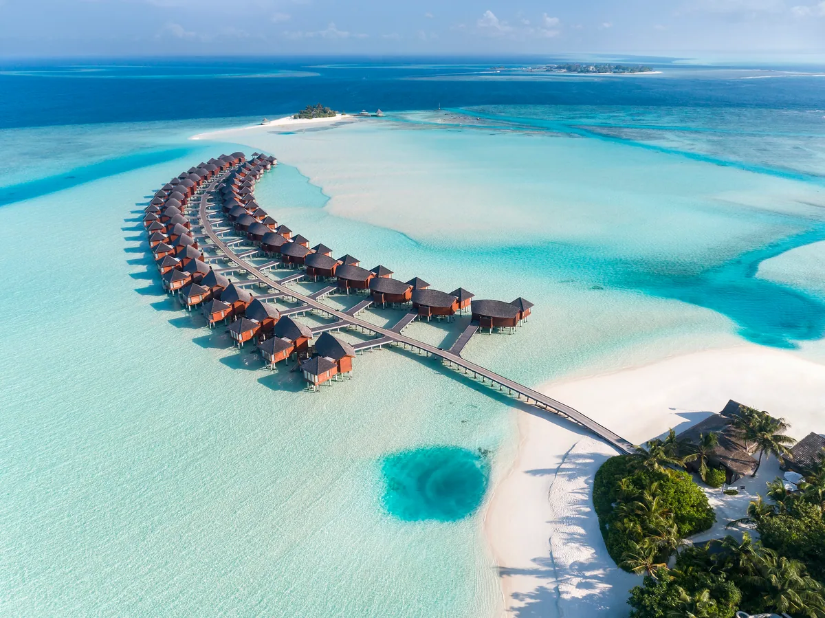 Anantara Dhigu Maldives Resort, aerial, South Malé Atoll, Maldives, Close aerial of the eastern over-water villa arc, the curving wing of red-shingle-roof Over-Water Villas extending from the white-sand beach island corner across the turquoise reef, the sand tongue at the villa cluster's base, and the adjacent Anantara Veli island visible at the top right of the frame.