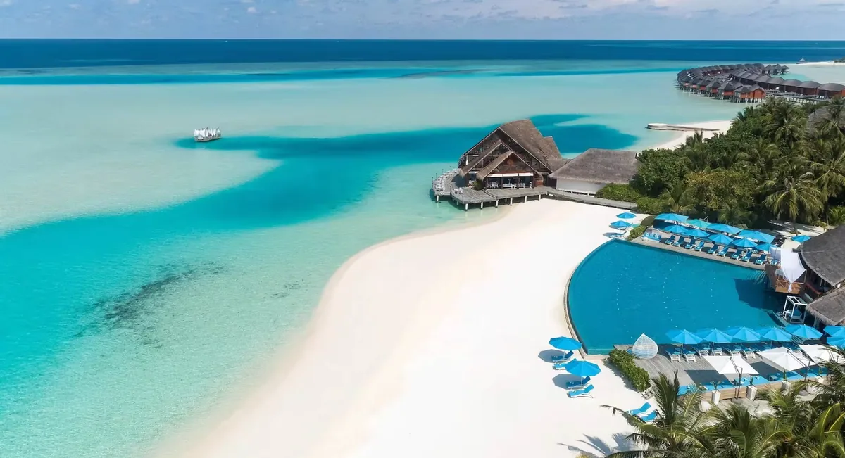 Anantara Dhigu Maldives Resort, setting, South Malé Atoll, Maldives, Beachfront infinity pool aerial, the large lagoon-edge pool with blue sun umbrellas in a radial arrangement and matching loungers, the open-walled lounge pavilion at the pool edge, the dhoni vessel anchored offshore against the reef, and the over-water villa row at the far right.