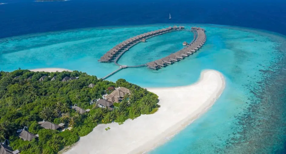 Anantara Kihavah Maldives Villas, aerial, Baa Atoll, Maldives, Kihavah Huravalhi from the east, the signature crescent-arc overwater villa cluster reaching off the central jetty, the curved sandbar wrapping the beach point, dense jungle interior, the far Baa Atoll island chain on the horizon.