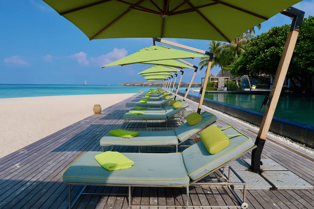 Anantara Kihavah Maldives Villas, pool, Baa Atoll, Maldives, Main pool deck, row of green-cushioned sun loungers under matching parasols on the teak boardwalk, infinity edge over the lagoon, the crescent overwater villa arc visible in the middle distance.
