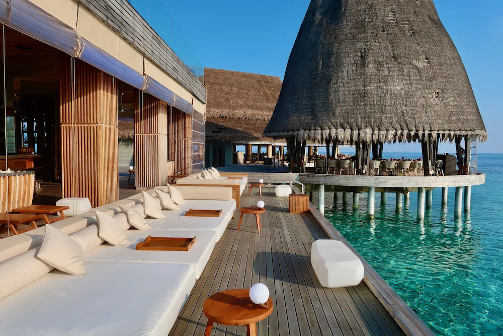 Anantara Kihavah Maldives Villas, bar, Baa Atoll, Maldives, Sky Bar overwater lounge deck: twin pointed conical thatched roofs (the Anantara Kihavah signature silhouette), expansive teak deck with low daybeds and white linen cushions, rope-wrapped pillars, glass-ball table lanterns, the lagoon edge to the right.