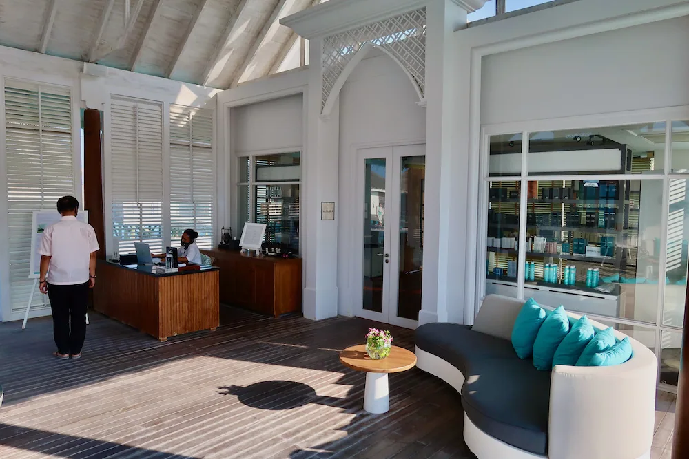 Anantara Kihavah Maldives Villas, spa, Baa Atoll, Maldives, Anantara Spa reception pavilion: vaulted white-painted ceiling with diamond fretwork detail above the gothic arch, louvered shutters, dark timber reception counter, curved teal-cushioned banquette, the retail dispensary visible through the glass partition.