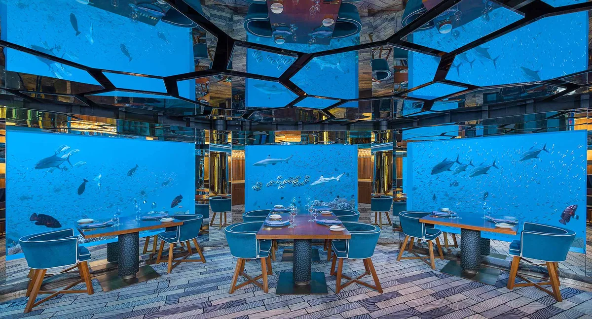 Anantara Kihavah Maldives Villas, dining, Baa Atoll, Maldives, SEA, the underwater restaurant four metres below the lagoon surface: four tables on a curved deck, walls and ceiling panelled in glass with reef life circulating beside the diners, mirrored ceiling reflecting the lagoon glow.