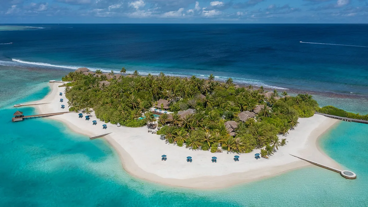 Naladhu Private Island Maldives, aerial, South Malé Atoll, Maldives, Elevated lateral aerial of the boutique island, white-sand crescent and shallow turquoise apron framing the dense palm canopy with the beach houses scattered under the trees; the timber walking jetty extends right onto the lagoon shelf and the boat jetty exits the western tip.