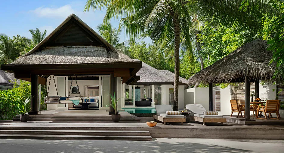 Naladhu Private Island Maldives, villa beach, South Malé Atoll, Maldives, Beach House beach-side overview, central thatched main pavilion with hanging-bed swing seat, plunge pool tucked behind the timber deck, separate thatched dining gazebo on the right with rattan armchairs, palm canopy framing the whole composition.