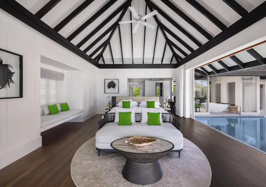Naladhu Private Island Maldives, villa beach, South Malé Atoll, Maldives, Jo Loves House bedroom, the Jo Malone scent-collaboration suite, colonial-style room with white panelled walls, black timber rafters under a pitched white ceiling, green accent pillows on the king bed, a circular ottoman with a carved-timber bowl, dark hardwood floor, plunge pool deck visible through full-height glass at right.