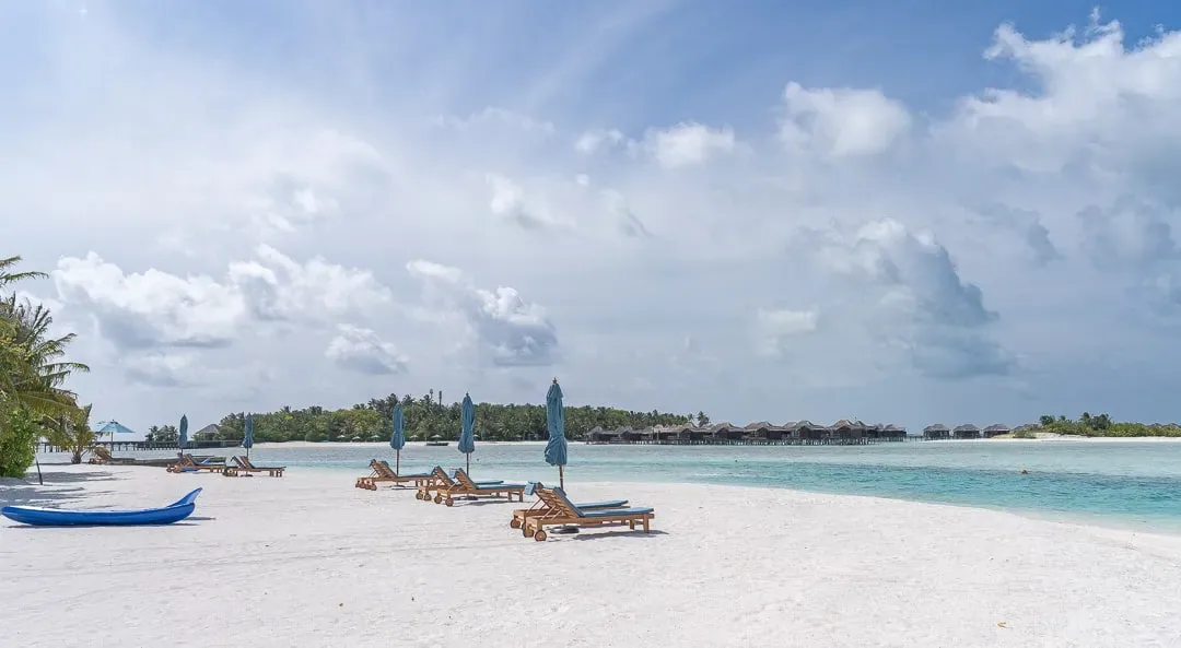 Naladhu Private Island Maldives, setting, South Malé Atoll, Maldives, Three-island cluster view from the Naladhu beach, white-sand strip with blue parasols and timber sun loungers in the foreground, the Anantara Veli over-water villa row visible across the cluster lagoon on the horizon line and a blue kayak pulled up on the sand.