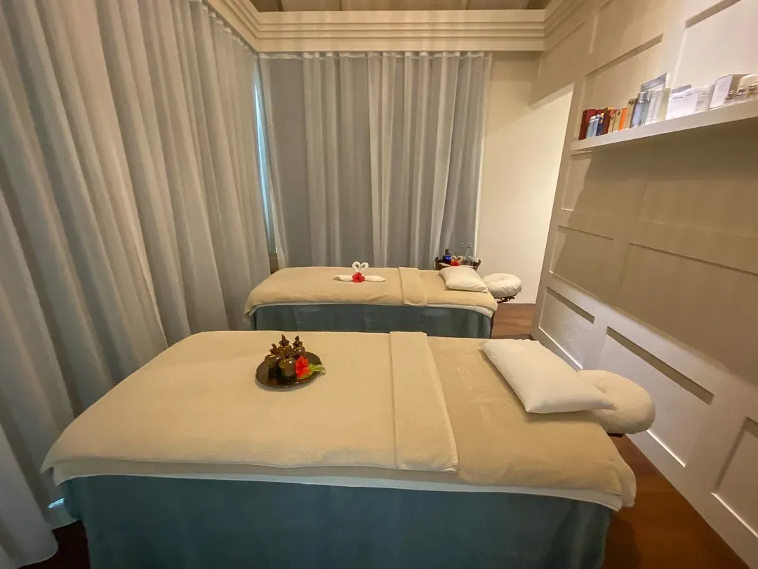 Naladhu Private Island Maldives, spa, South Malé Atoll, Maldives, Naladhu spa twin treatment room, beige-and-blue palette with full-length sheer curtains around the perimeter, two side-by-side massage beds with white-towel arrangements, brass essential-oil tray and red flower petals between, white panelled feature wall right with display shelves.
