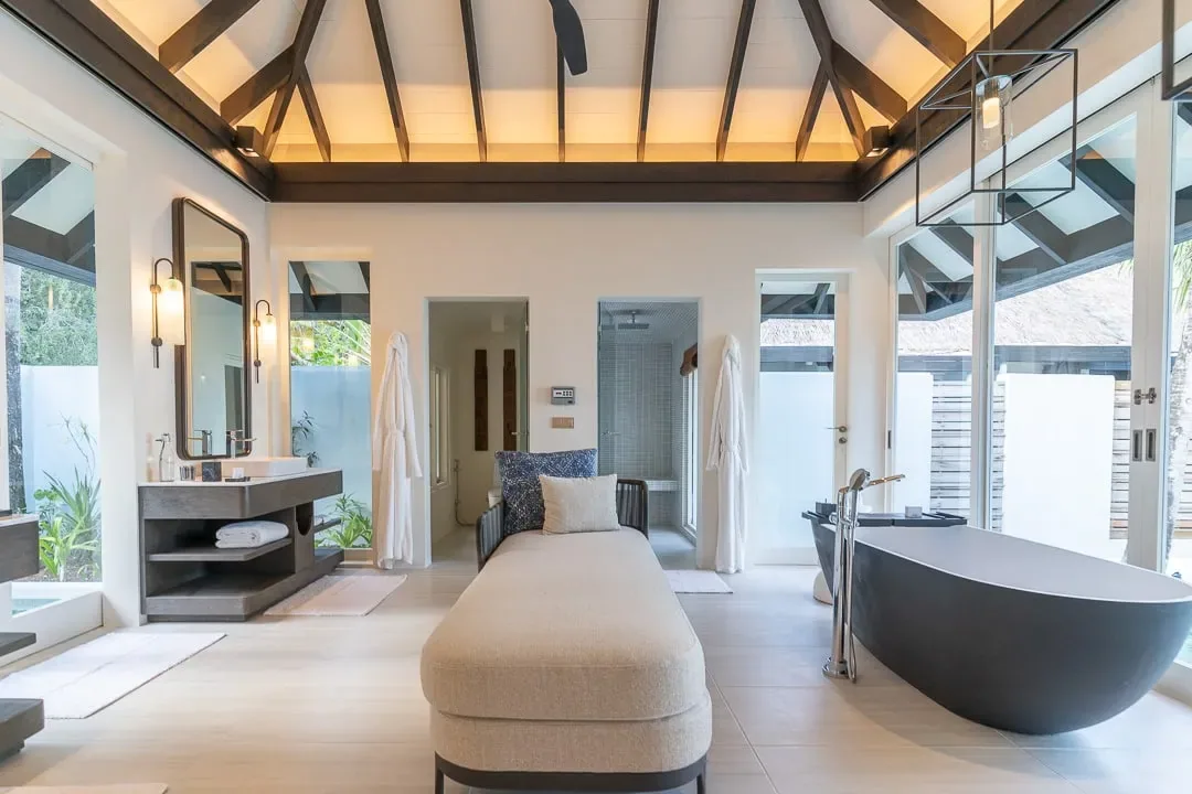 Naladhu Private Island Maldives, villa beach, South Malé Atoll, Maldives, Beach House bathroom, dark timber pitched ceiling with exposed rafters, twin vanities on the left, freestanding charcoal-grey oval tub on the right with a chrome floor-mount faucet, central beige chaise, full-height glass on three sides opening to private courtyards.