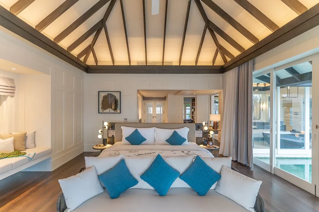 Naladhu Private Island Maldives, villa beach, South Malé Atoll, Maldives, Beach House bedroom interior, pitched white ceiling with dark timber rafters, white-shutter panelling, four-poster bed with a flower-petal welcome message and teal accent pillows, daybed at the foot of the bed, window-seat reading nook on the left, plunge pool visible through glass doors on the right.