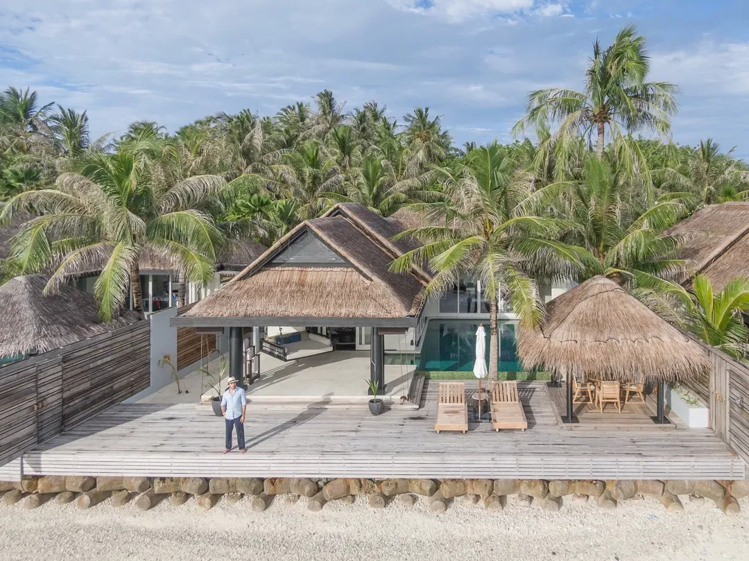 Naladhu Private Island Maldives, villa beach, South Malé Atoll, Maldives, Ocean House front exterior from the beach, two thatched gable-roofed pavilions linked by a covered deck, palm-canopy backdrop, staff member in white walking the deck for scale.