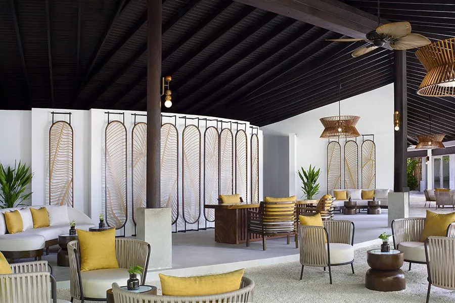 Anantara Veli Maldives Resort, arrival, South Malé Atoll, Maldives, Reception pavilion post-2022 Yamazaki renovation, black-stained timber pitched ceiling with exposed beams, vertical white screens with palm-leaf cutouts, woven cane pendant lights and mustard accent cushions, the contemporary-Japanese restraint that replaced the prior tropical-luxury saturation.
