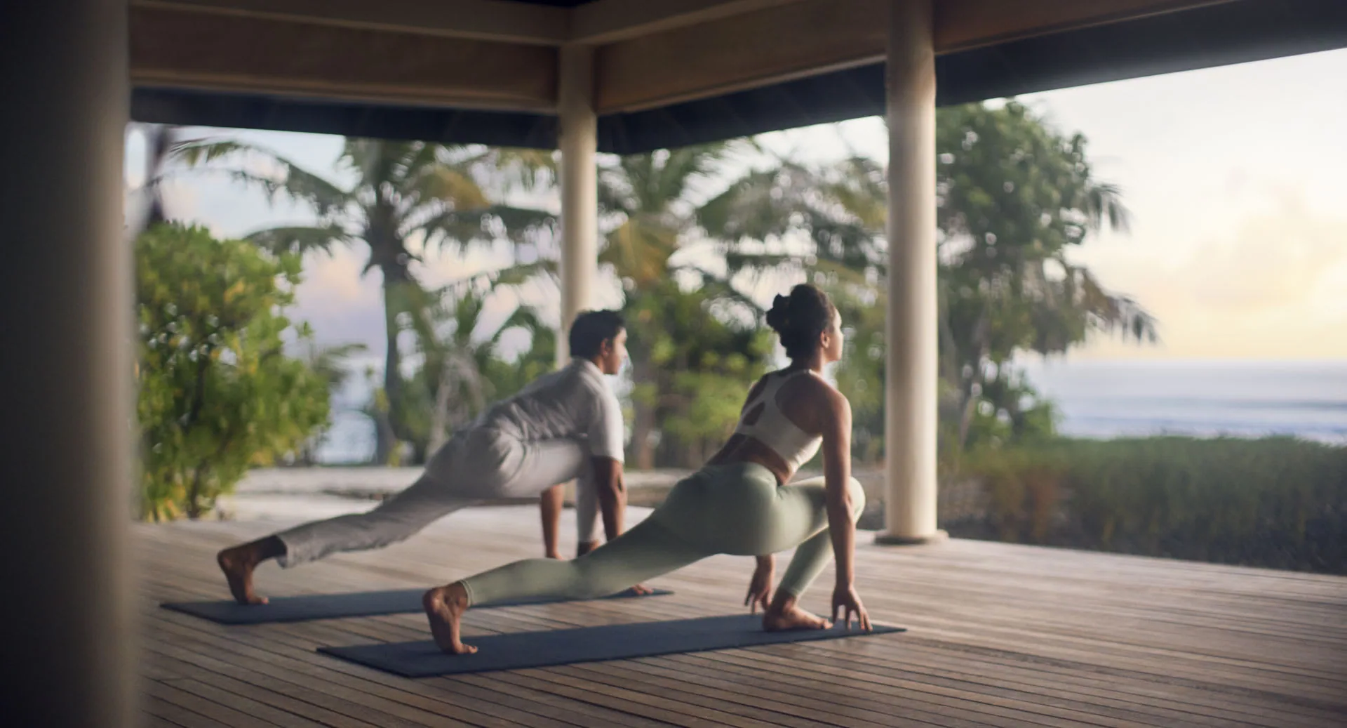Anantara Veli Maldives Resort, activities, South Malé Atoll, Maldives, Sunrise yoga session at the Wellness Villa pavilion, two practitioners in low lunge on the hardwood deck, palm trees and the ocean horizon visible beyond the open eave.