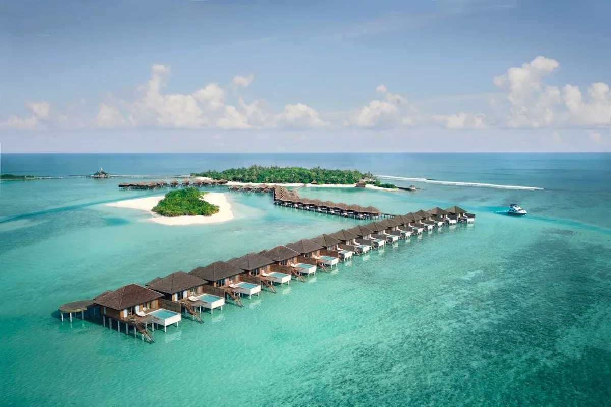 Anantara Veli Maldives Resort, setting, South Malé Atoll, Maldives, Veligandu Huraa island aerial, the linear cluster of overwater pool villas extending into the eastern lagoon, small palm-fringed sandbank in the foreground and the property's protected reef visible through the calm surface.