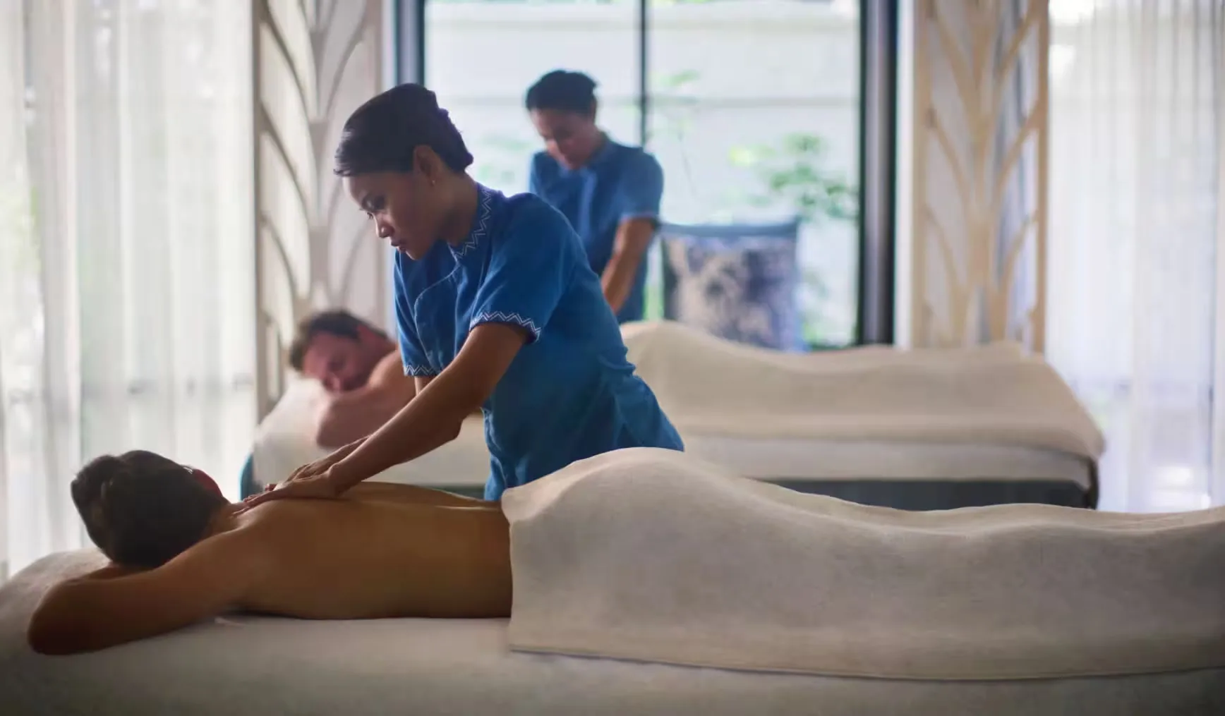 Anantara Veli Maldives Resort, spa, South Malé Atoll, Maldives, Couples treatment in the post-renovation spa, twin massage beds with Anantara therapists in blue uniforms, soft sheer curtains and the Yamazaki vertical-screen detailing in the background.