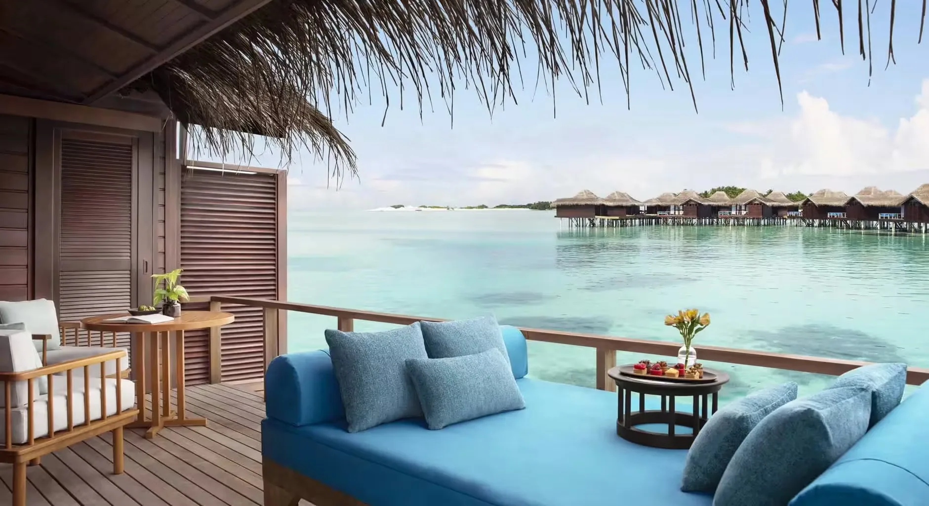 Anantara Veli Maldives Resort, villa overwater, South Malé Atoll, Maldives, Over Water Villa deck after the 2022 Yamazaki renovation, woven thatched eave, sun-bleached timber decking with a blue-cushioned daybed and side chair, view across the lagoon to the linear villa cluster and sandbank.