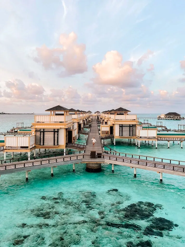 Angsana Velavaru, villa overwater, Dhaalu Atoll, Maldives, The signature InOcean Villa cluster viewed from the central timber boardwalk at sunset, receding rows of two-storey yellow-walled villas with rooftop terraces and grey-tiled sloped roofs on both sides, a guest walking down the central boardwalk over the lagoon reef.