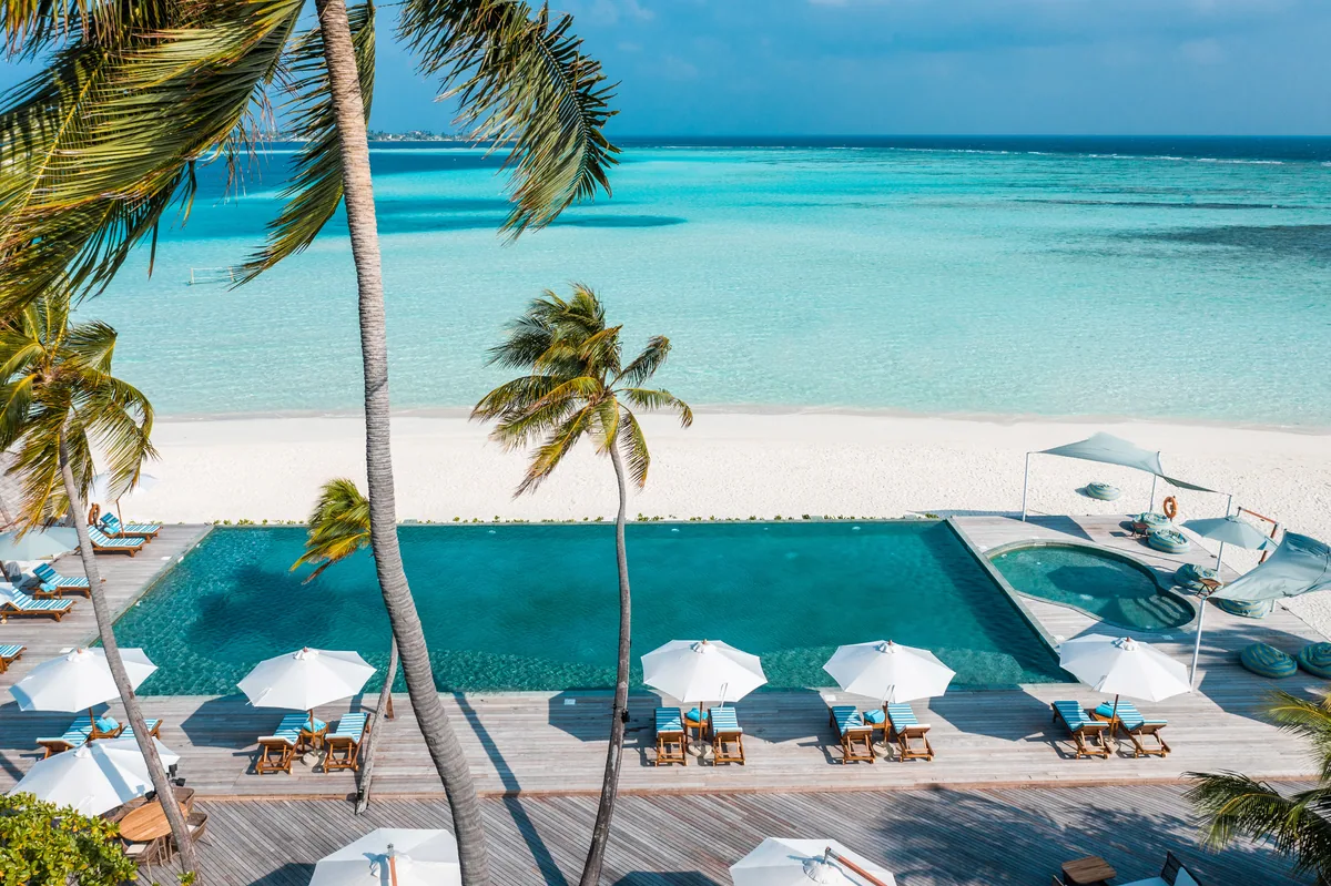 Angsana Velavaru, dining, Dhaalu Atoll, Maldives, Main-island infinity pool with white umbrellas and symmetric loungers along the timber deck, palm trees framing the left edge, white-sand beach in middle ground, the lagoon and distant island on the horizon.