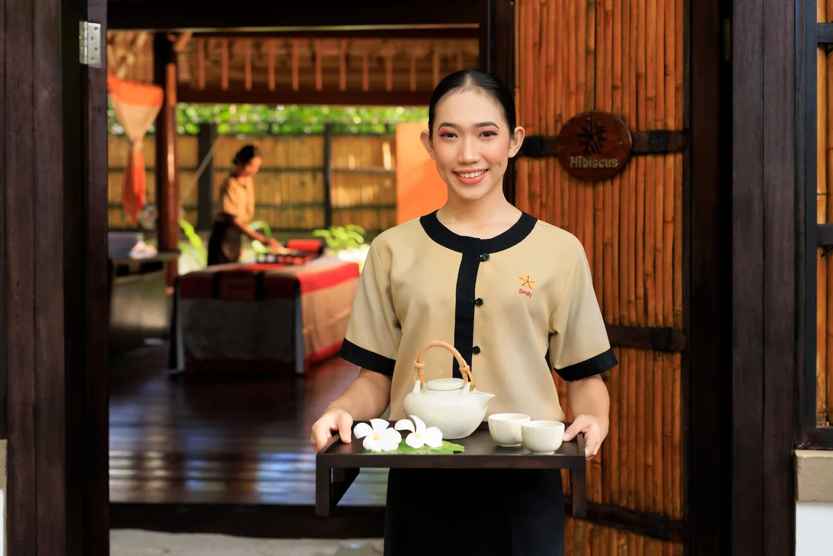 Angsana Velavaru, spa, Dhaalu Atoll, Maldives, Spa welcome at the Hibiscus treatment cabin, a therapist with a tea-and-plumeria tray standing in the wood-framed cabin doorway with the carved Hibiscus plaque, treatment bed visible inside the cabin.