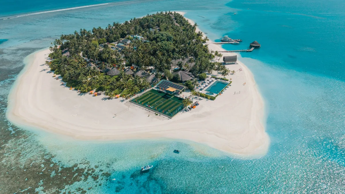 Angsana Velavaru, aerial, Dhaalu Atoll, Maldives, High-angle aerial of the main Velavaru island showing the activity cluster: futsal court, infinity pool, palm-canopy interior, beach-villa cluster, jetty walkway leading out toward the InOcean platform, and the outermost over-water villa pavilions visible at right.