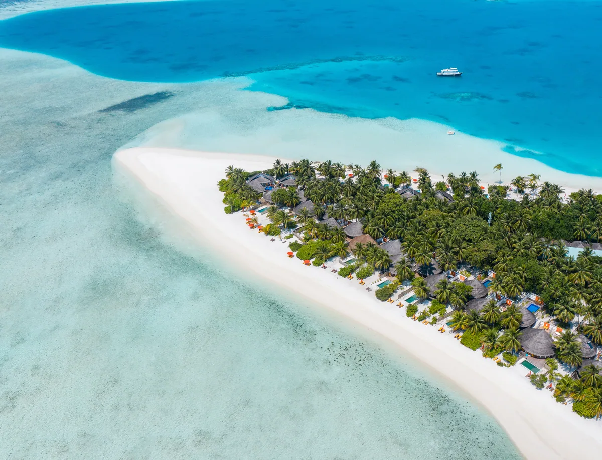 Angsana Velavaru, aerial, Dhaalu Atoll, Maldives, Aerial of the southwest Velavaru island tip with the white-sand sandspit extending into the lagoon, beach-villa cluster beneath the palm canopy, dive boat moored offshore in the deep reef.