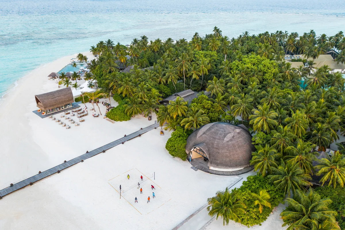 Angsana Velavaru, aerial, Dhaalu Atoll, Maldives, Oblique aerial of the activity area on the main island, distinctive curved shell-roof building (the architectural signature) beside a sand-floor beach volleyball court, jetty walkway extending out, dive-boat slip and orange umbrellas along the beach line.