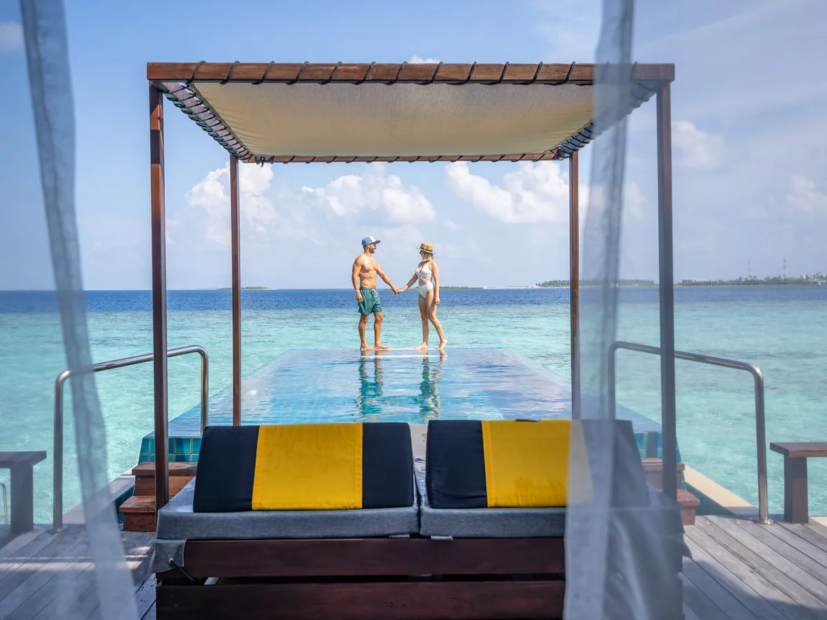 Angsana Velavaru, villa overwater, Dhaalu Atoll, Maldives, InOcean villa cantilevered infinity pool viewed from the villa interior, a couple at the far end of the pool framed by sheer balcony curtains, signature yellow-and-black striped daybed cushion in the foreground, open-ocean view to the horizon.