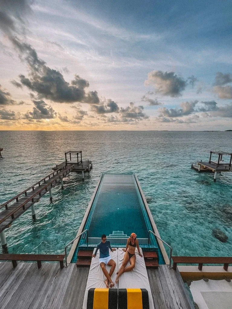 Angsana Velavaru, villa overwater, Dhaalu Atoll, Maldives, Rooftop terrace at golden hour, the signature yellow-and-black striped daybed with a couple watching the sunset, cantilevered infinity pool stretching toward the horizon, the two neighboring villa rooftop pavilions framing the composition at left and right.