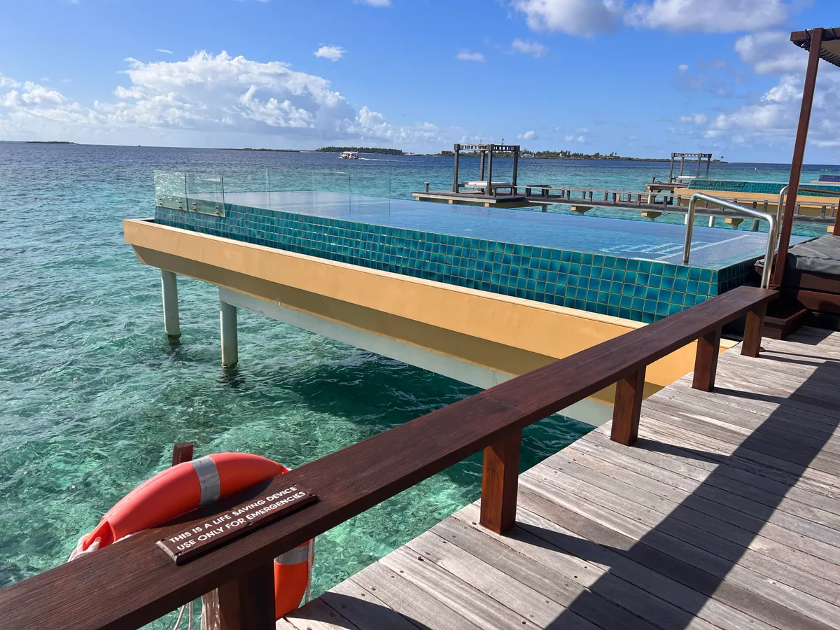 Angsana Velavaru, villa overwater, Dhaalu Atoll, Maldives, Cantilevered InOcean pool viewed from the side deck, the signature yellow underside and green-tiled outer wall (the property's architectural signature), glass infinity-edge panels, life ring on the rail, view across to neighboring villa pavilions and the main island.