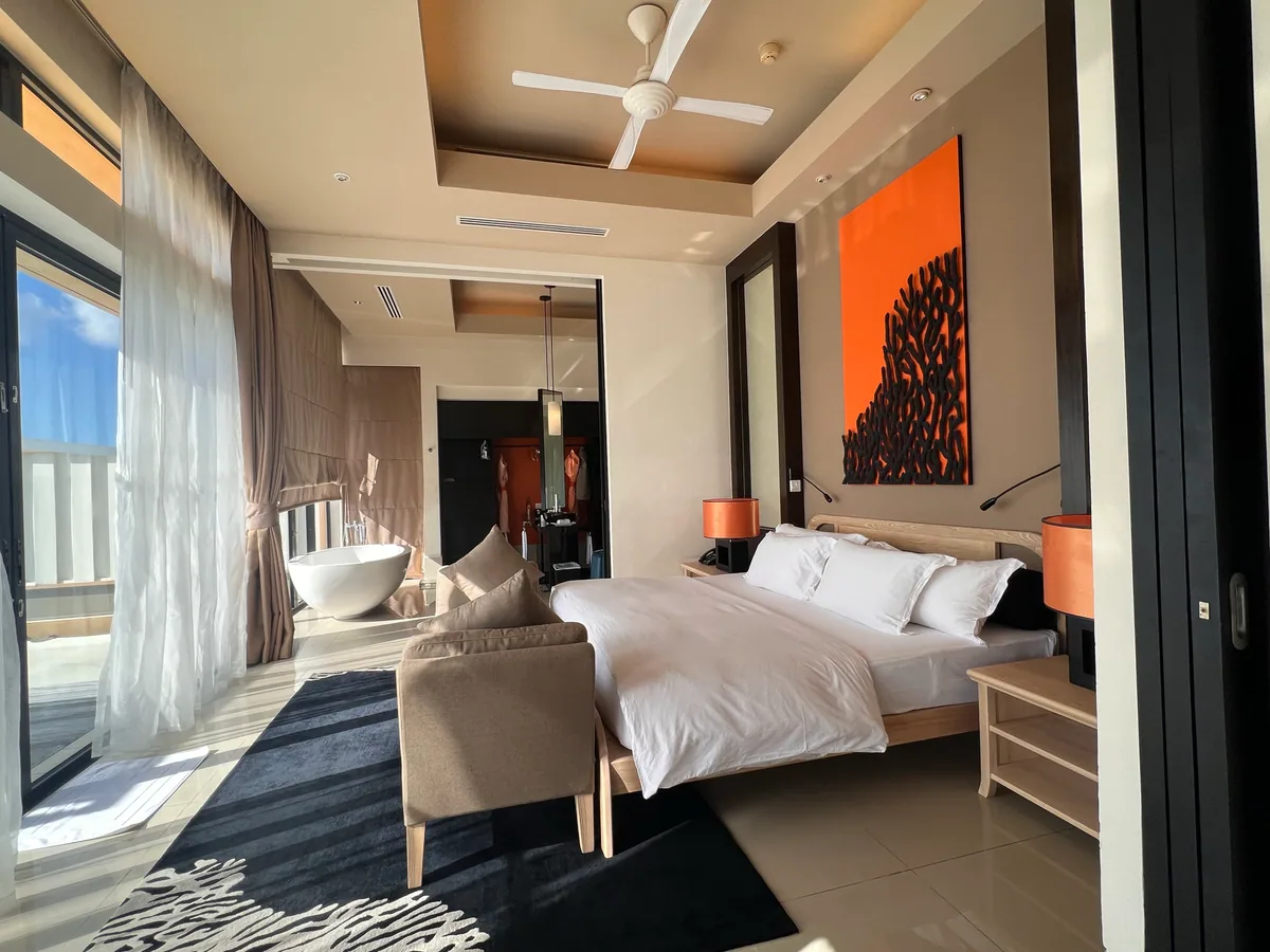 Angsana Velavaru, villa interior, Dhaalu Atoll, Maldives, InOcean villa bedroom, king bed with white linens beneath a large orange-on-black coral-motif canvas (the brand's signature interior detail), egg-shaped freestanding soaking tub at the bedroom edge, beige modular sofa, sheer balcony curtains opening to the deck.