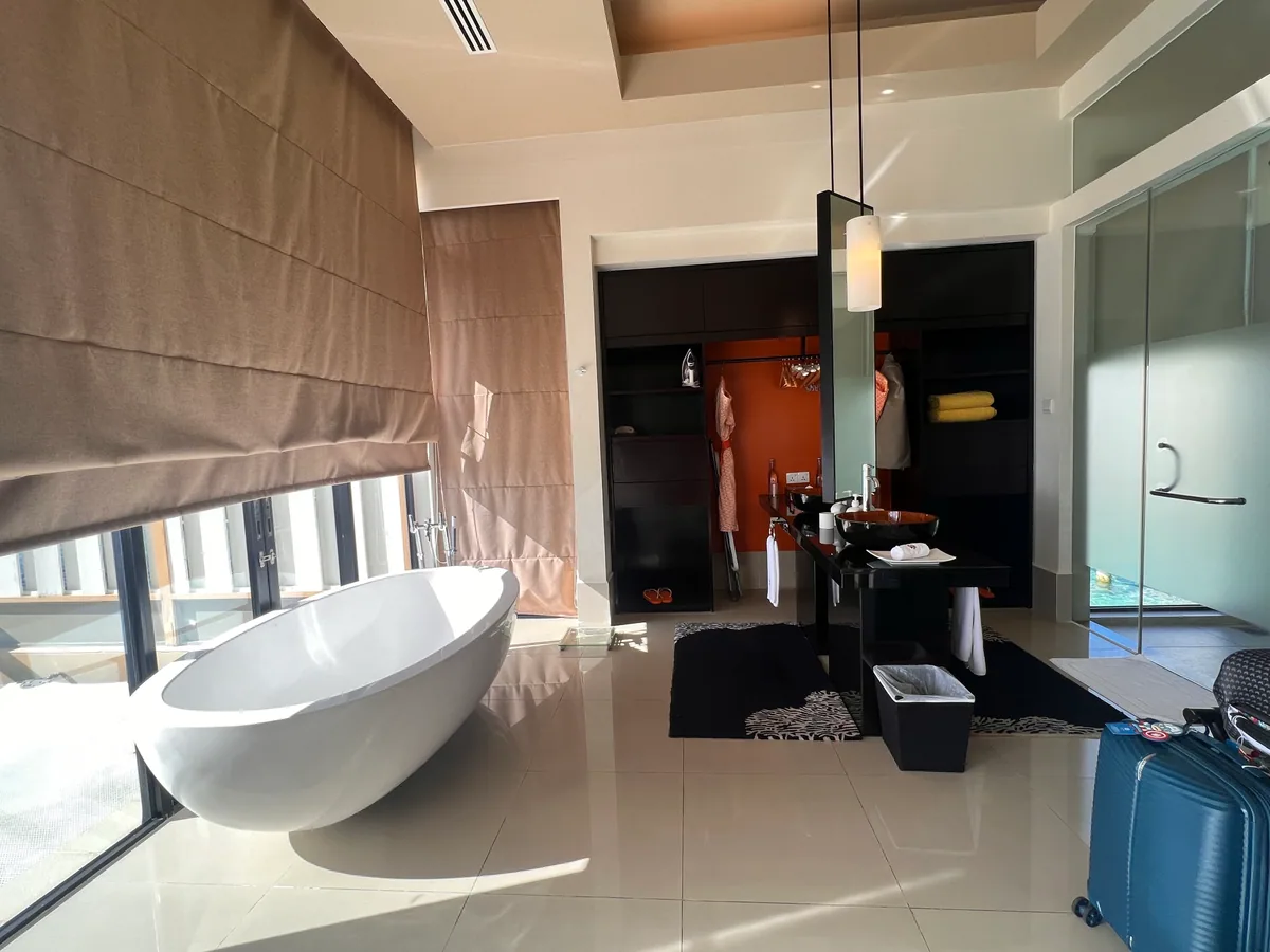Angsana Velavaru, villa interior, Dhaalu Atoll, Maldives, InOcean villa bathroom, egg-shaped white freestanding tub at the picture-frame window, dark vanity with vessel sink, walk-in wardrobe with the brand's orange feature wall (Angsana signature) at the back, glass shower enclosure at right.