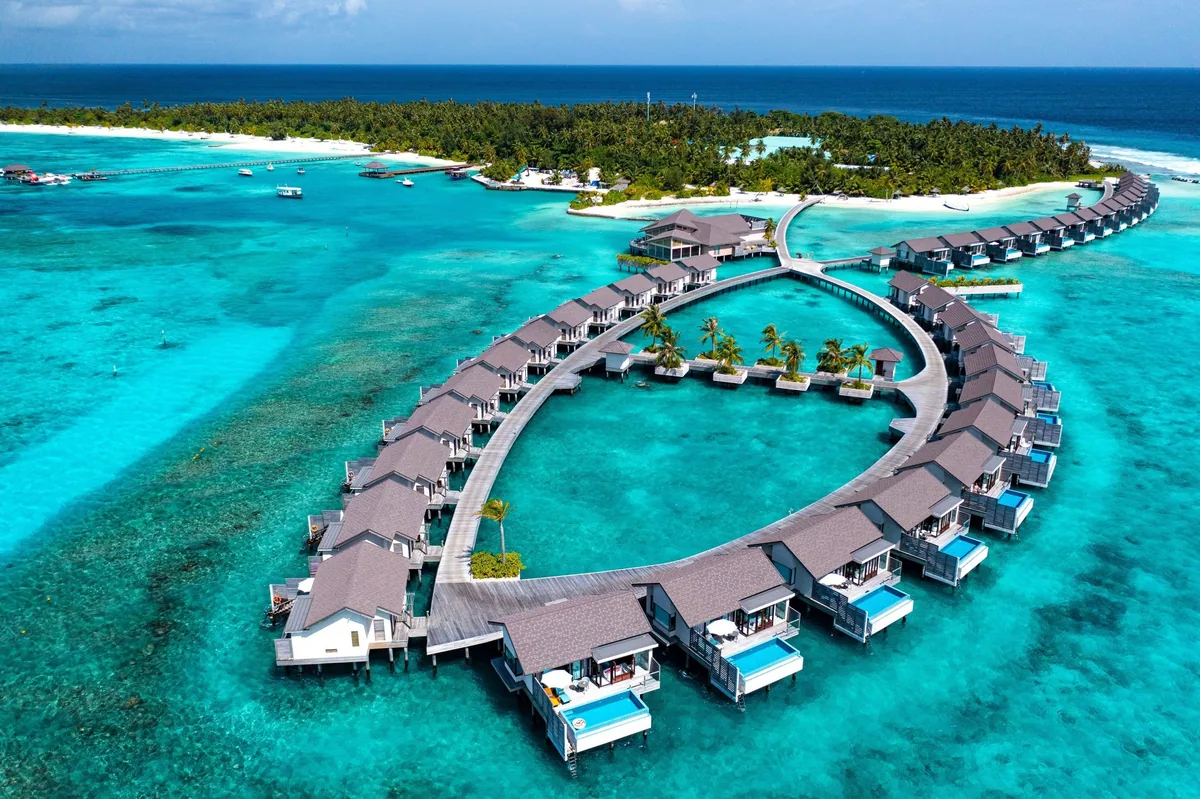 Atmosphere Kanifushi Maldives, aerial, Lhaviyani Atoll, Maldives, Curved ring of forty overwater villas with the Pier Six restaurant peninsula at the centre, looking back toward the longer-axis Kanifushi beach island.