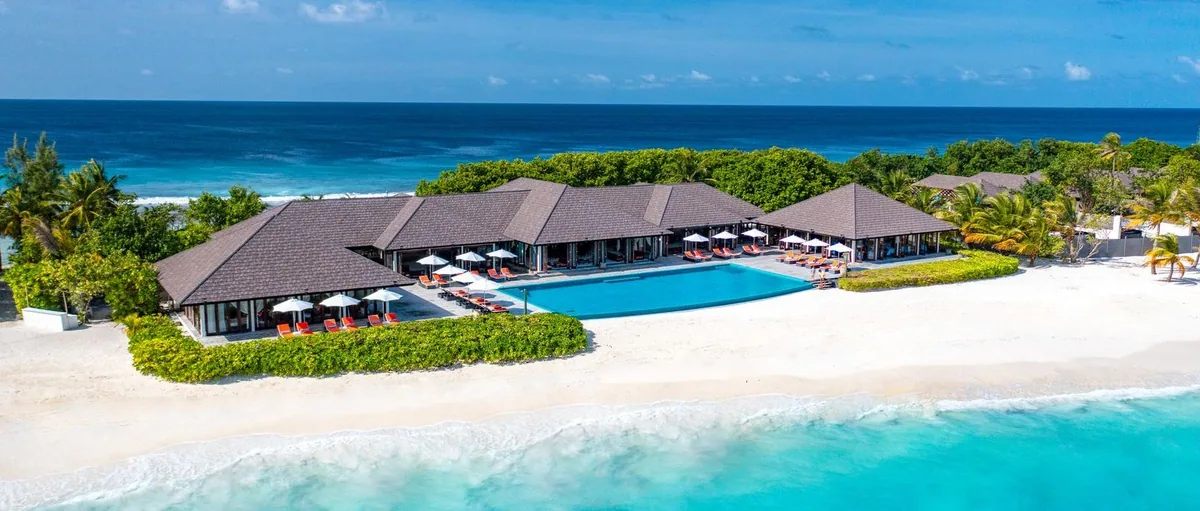 Atmosphere Kanifushi Maldives, aerial, Lhaviyani Atoll, Maldives, Mid-elevation view of the central restaurant pavilion cluster with the pool, orange sun loungers, and the reef break beyond the beach.