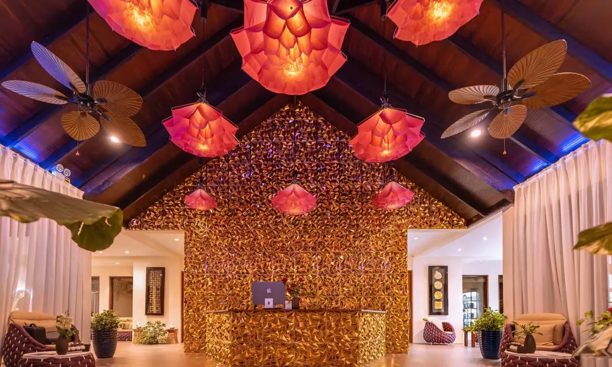 Atmosphere Kanifushi Maldives, spa, Lhaviyani Atoll, Maldives, Essens Spa lobby with a golden mosaic feature wall, coral-petal pendant lights, palm-frond ceiling fans, and the reception desk centred under the vault.