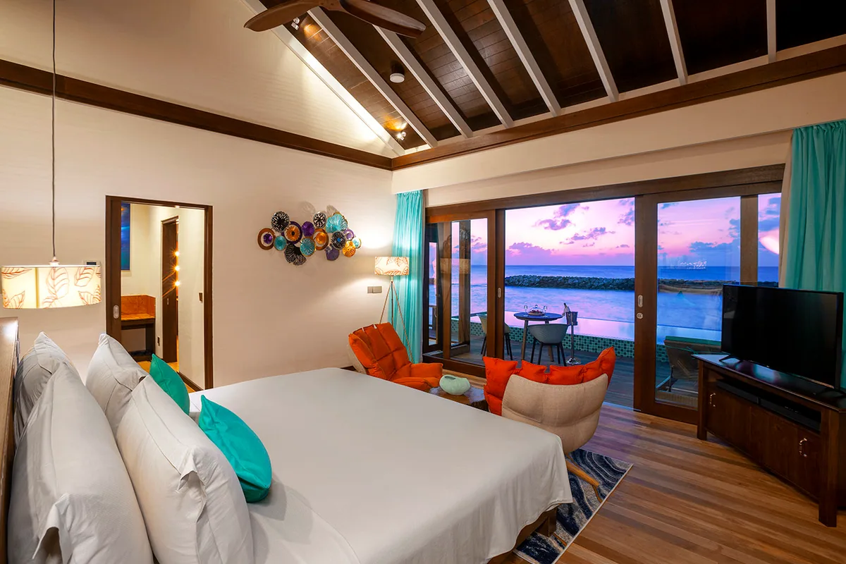 Atmosphere Kanifushi Maldives, villa overwater, Lhaviyani Atoll, Maldives, Sunset Water Villa with Pool bedroom at dusk, vaulted timber ceiling, decorative ceramic-plate wall mural, teal accents, and sliding doors to the private overwater deck.