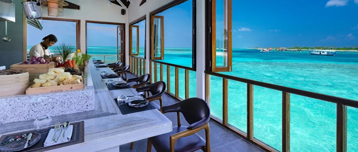 Atmosphere Kanifushi Maldives, dining, Lhaviyani Atoll, Maldives, Pier Six overwater restaurant counter with a chef prepping vegetables, place settings facing the lagoon, and a seaplane on the distant reef.