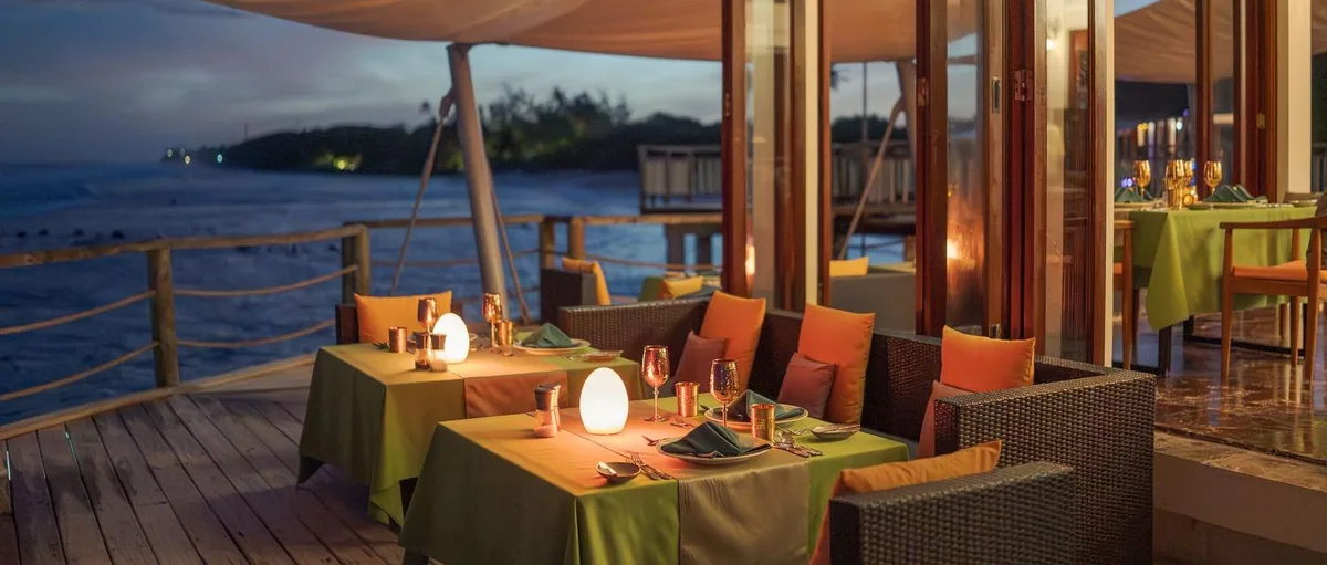 Atmosphere Kanifushi Maldives, dining, Lhaviyani Atoll, Maldives, Just Veg overwater dining deck at dusk, green linens with copper Indian-themed glassware, egg-candle lanterns, and a sail-canopy overhead.