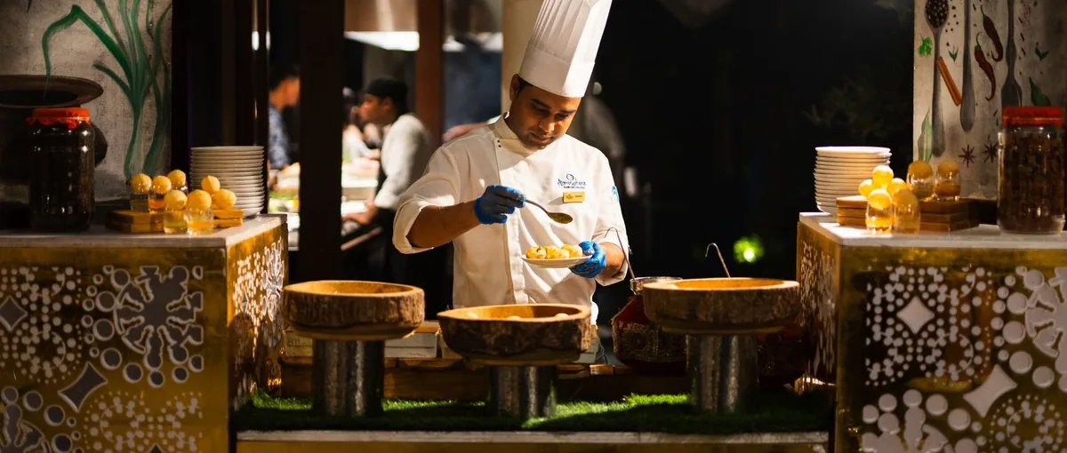 Atmosphere Kanifushi Maldives, dining, Lhaviyani Atoll, Maldives, Spice principal all-day pavilion with a chef plating saffron-coloured dishes at the live station, gold-mosaic counter, and a vegetable-and-spice mural backdrop.
