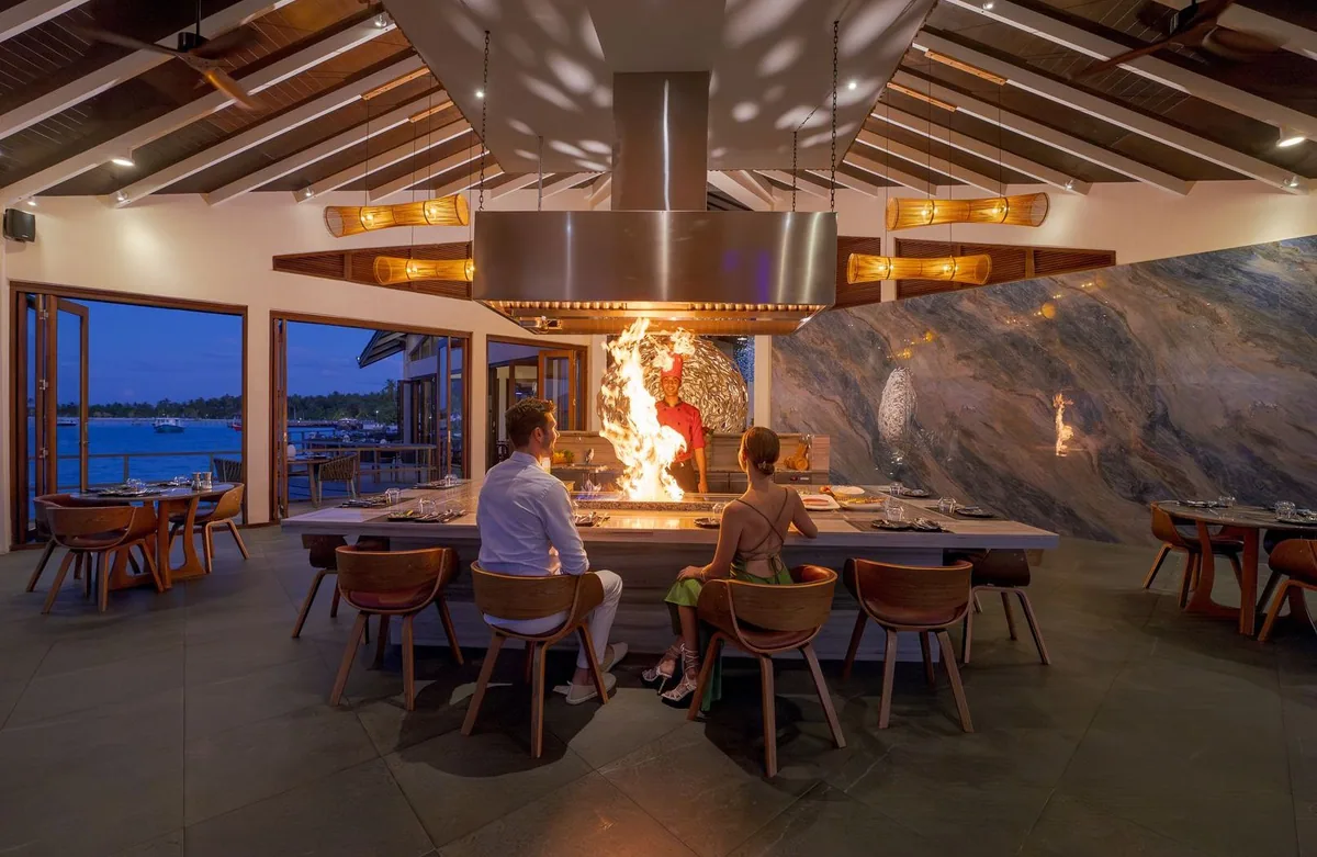 Atmosphere Kanifushi Maldives, dining, Lhaviyani Atoll, Maldives, Teppanyaki Grill counter with a chef and live flame, vaulted ceiling and cone pendant lights, marble feature wall, and lagoon dusk view through opened doors.