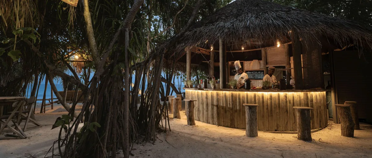 Atmosphere Kanifushi Maldives, dining, Lhaviyani Atoll, Maldives, Ceylon Bliss Sri Lankan-themed beach bar built into the pandanus trees, semi-circular wooden counter with under-counter lighting, and log stools on sand.