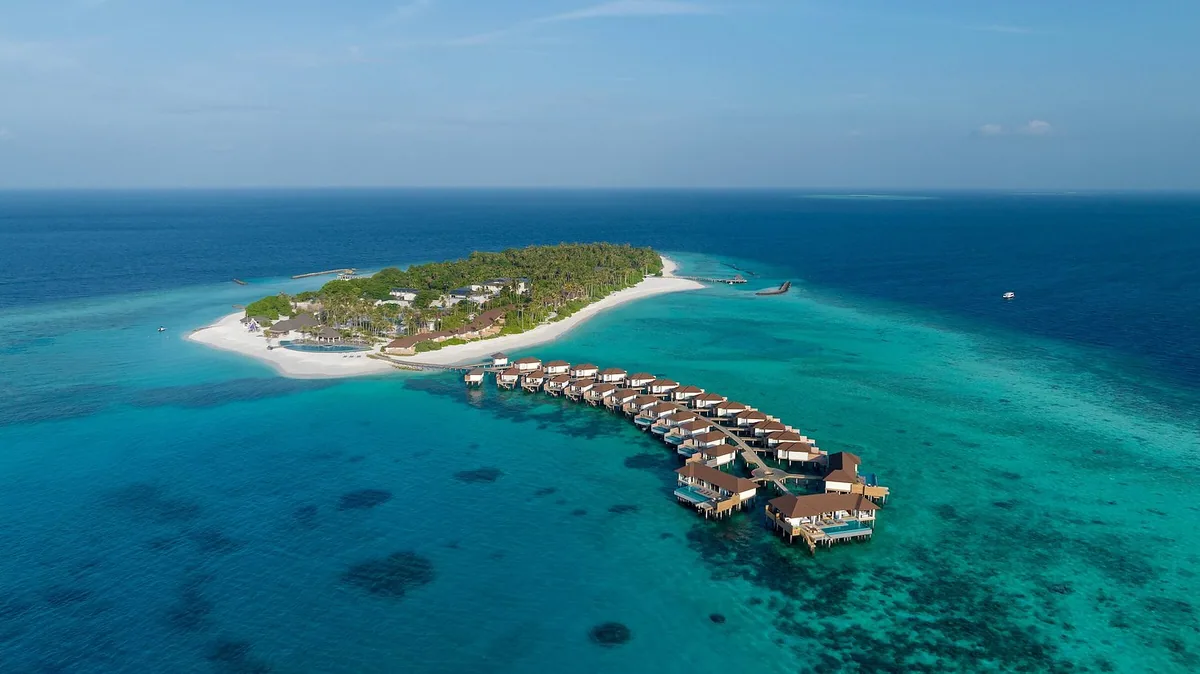 Avani+ Fares Maldives, Fares island aerial with palm canopy, central beach pool, curved water villa cluster extending from jetty on turquoise lagoon, Baa Atoll, Maldives