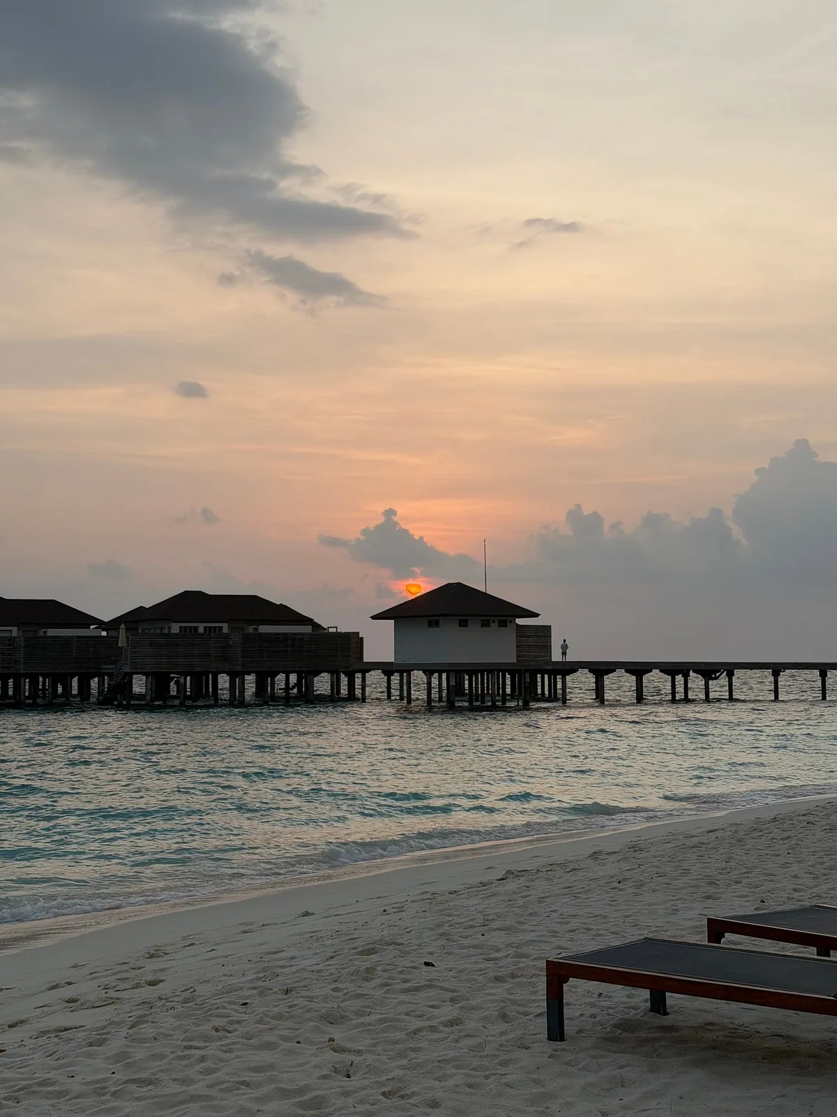 Avani+ Fares Maldives, sunset over water villa jetty with pitched-roof villas, central jetty-end pavilion and beach sun loungers in foreground, Baa Atoll, Maldives