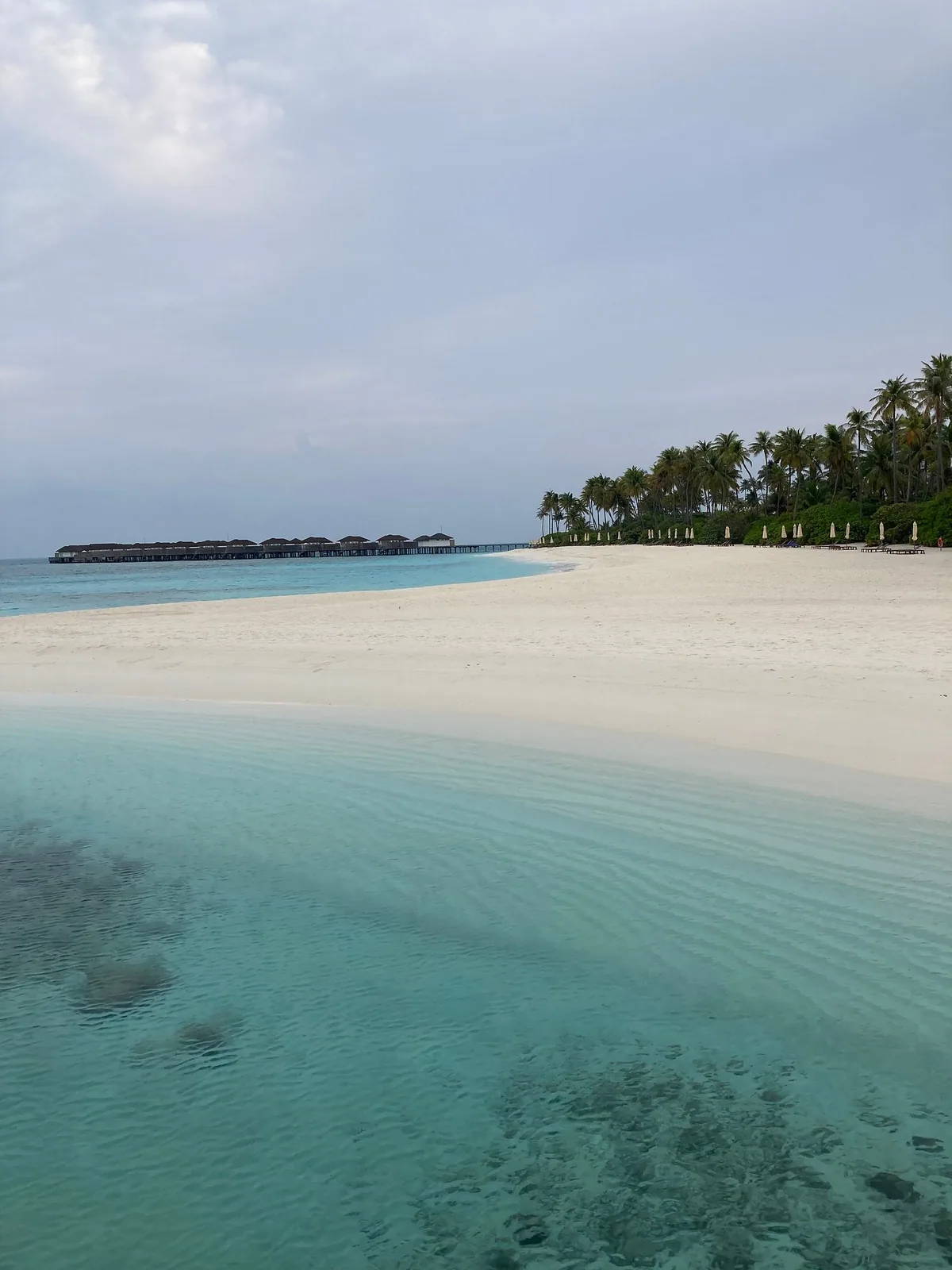 Avani+ Fares Maldives, white-sand spit shallows with water villa jetty cluster on horizon left and palm coastline with beach loungers right, Baa Atoll, Maldives