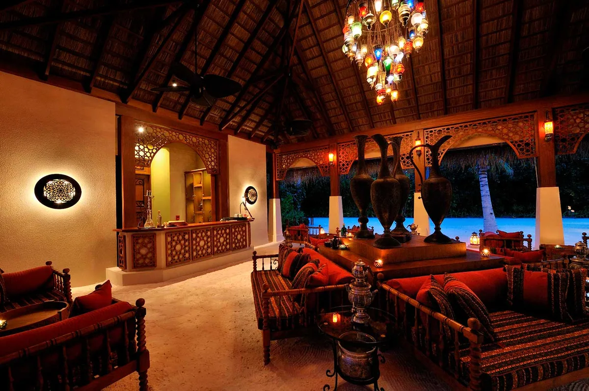 Ayada Maldives, dining, Gaafu Dhaalu Atoll, Maldives, Ottoman Lounge interior at dusk: stained-glass Turkish chandelier suspended from thatched cathedral ceiling, ornate fretwork wood screens, antique Ottoman brass jugs grouped on plinth, kilim-pattern cushions on dark hardwood divans, hookah pipes ready beside low brass tables. The Turkish character rendered in furniture.