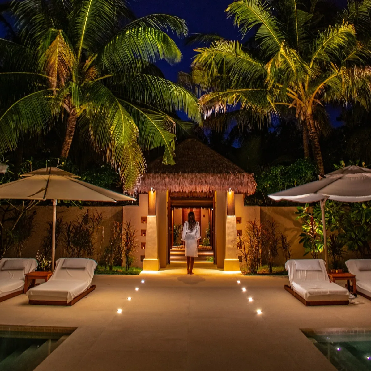 Ayada Maldives, spa, Gaafu Dhaalu Atoll, Maldives, AySpa entrance pavilion at night: thatched-roof spa entry flanked by twin plunge pools, terraced loungers under matching umbrellas, soft ground-lighting tracing the causeway, a guest in white spa robe walking the spine path into the treatment cluster.