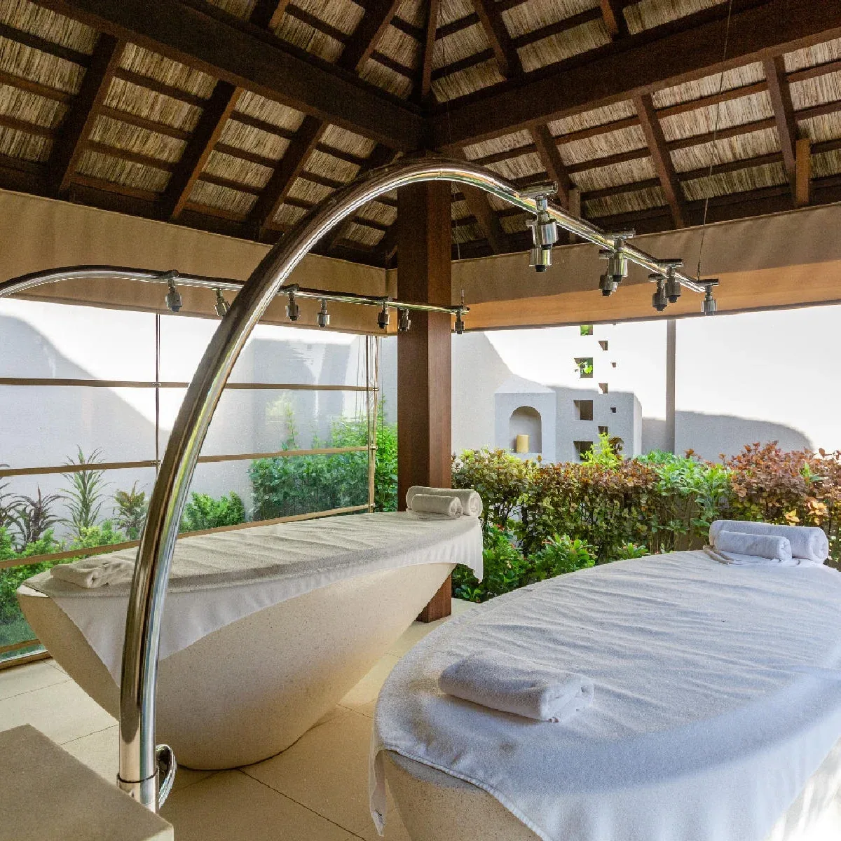 Ayada Maldives, spa, Gaafu Dhaalu Atoll, Maldives, AySpa treatment villa interior: twin parallel massage beds under a thatched pitched ceiling, vichy-shower arc rail mounted over one bed, open-air courtyard with planted hedge wall and second pavilion visible through the screen.