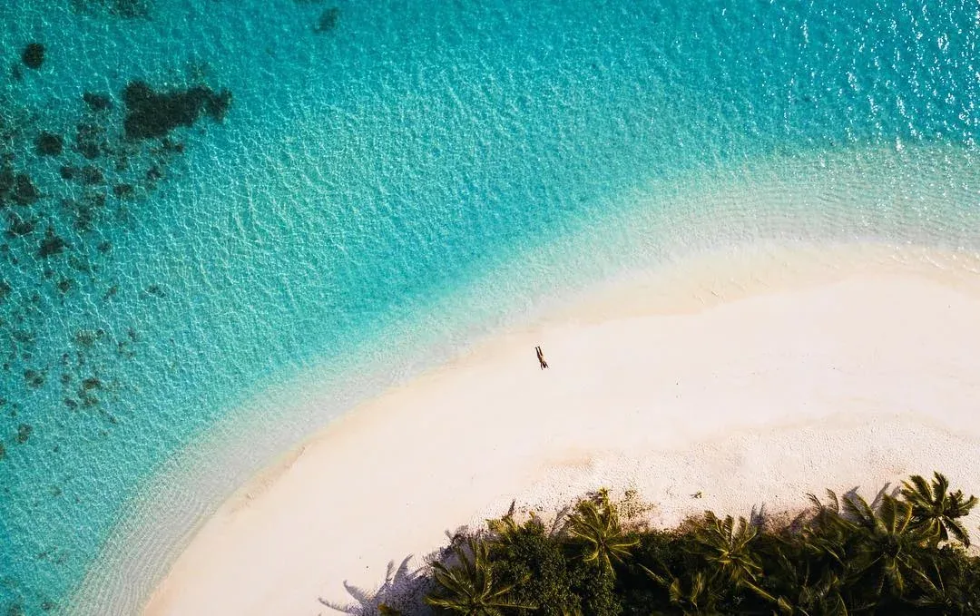 Ayada Maldives, setting, Gaafu Dhaalu Atoll, Maldives, Aerial of the Maguhdhuvaa beach perimeter at midday, turquoise lagoon shading to navy over the reef drop, lone guest walking the white-sand tip near the palm fringe, house-reef coral structure visible underwater.