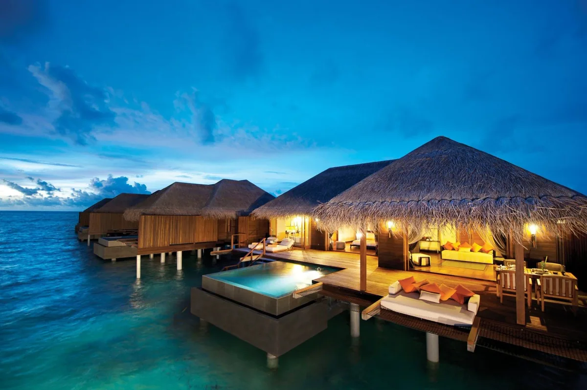 Ayada Maldives, villa overwater, Gaafu Dhaalu Atoll, Maldives, Sunset Ocean Suite cluster at blue hour, thatched-pyramid roofs above stilted overwater villas, private plunge pool stepped into the deck, saffron-orange daybed cushions (Ottoman colour palette) against bleached teak boards, lagoon water still as glass.