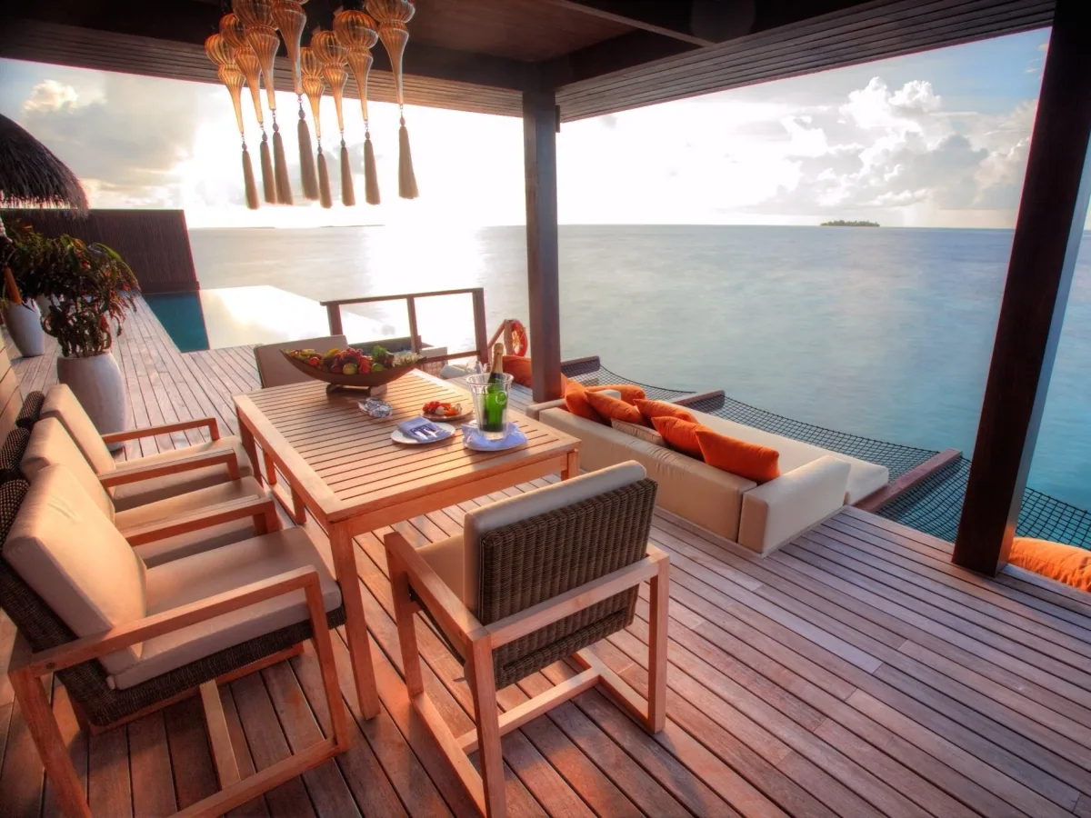 Ayada Maldives, villa overwater, Gaafu Dhaalu Atoll, Maldives, Royal Ocean Suite open-air deck, silver-tassel Turkish pendant lights suspended over the dining table, low corner sofa with saffron cushions, infinity-edge plunge pool stepping into the lagoon, distant island silhouette on the horizon.